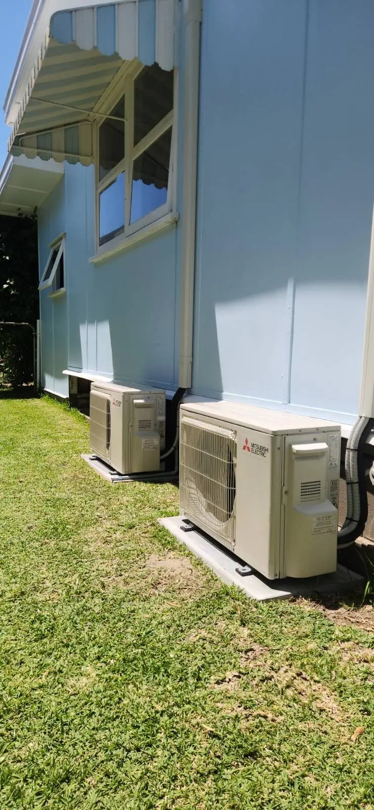Two Mitsubishi Electric air conditioning outdoor units on concrete pads beside a pale blue weatherboard house