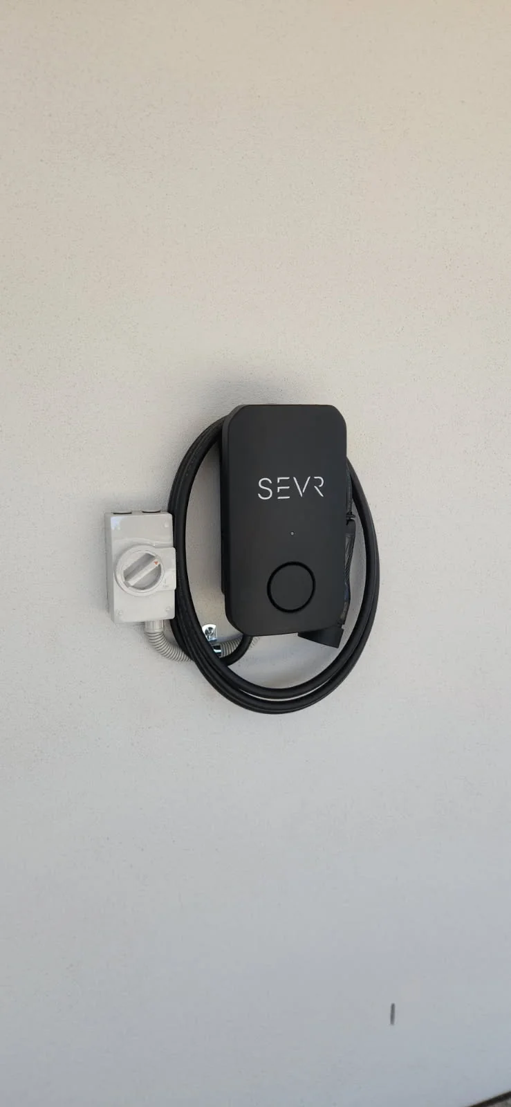 Black SEVR electric vehicle charger with a coiled cable and an isolator switch on a rendered wall